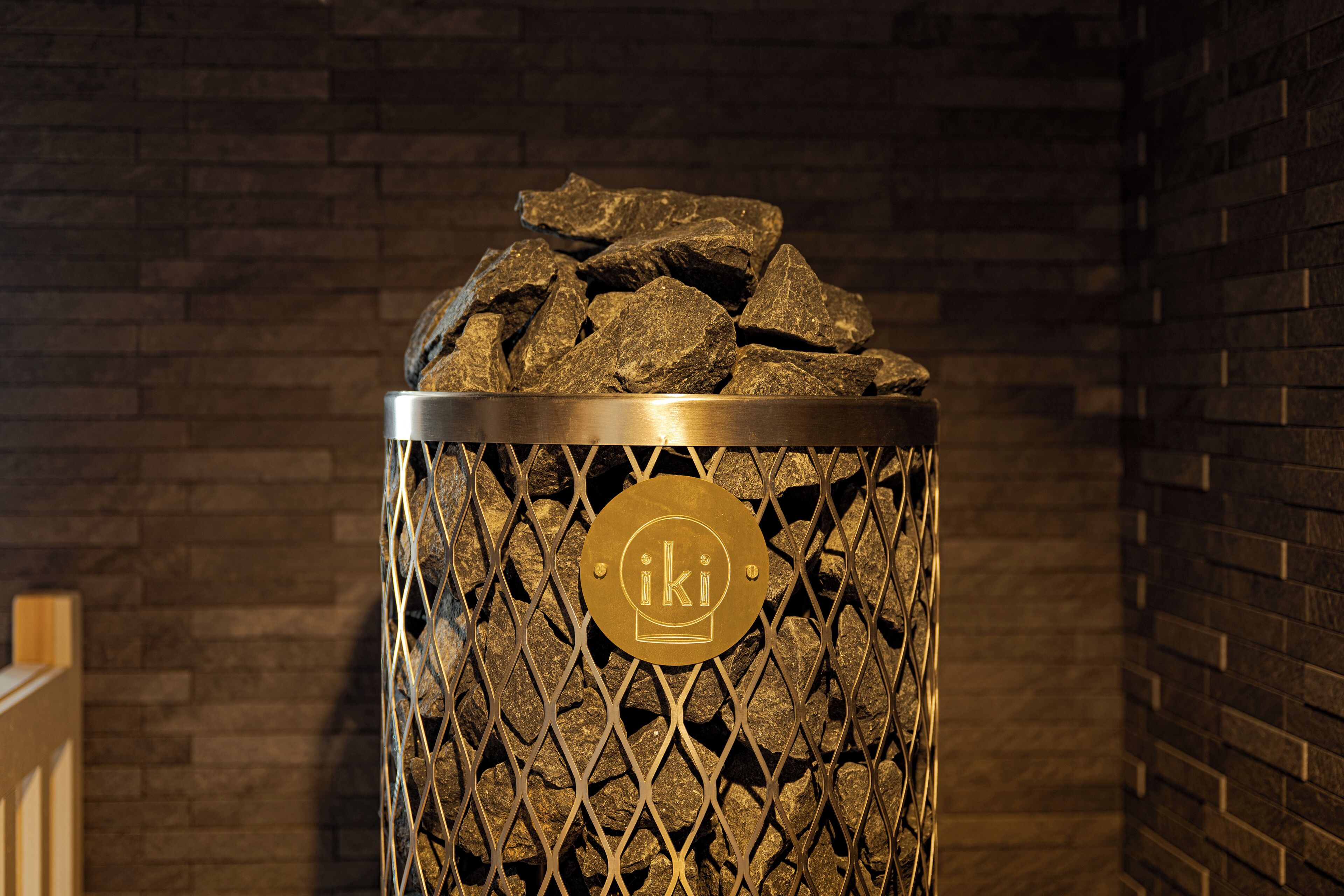 A metal trash bin with a decorative metal mesh and a circular emblem, filled with rocks, against a brick wall background.