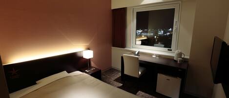 A hotel room with a bed, desk, and a window with a city view at night.