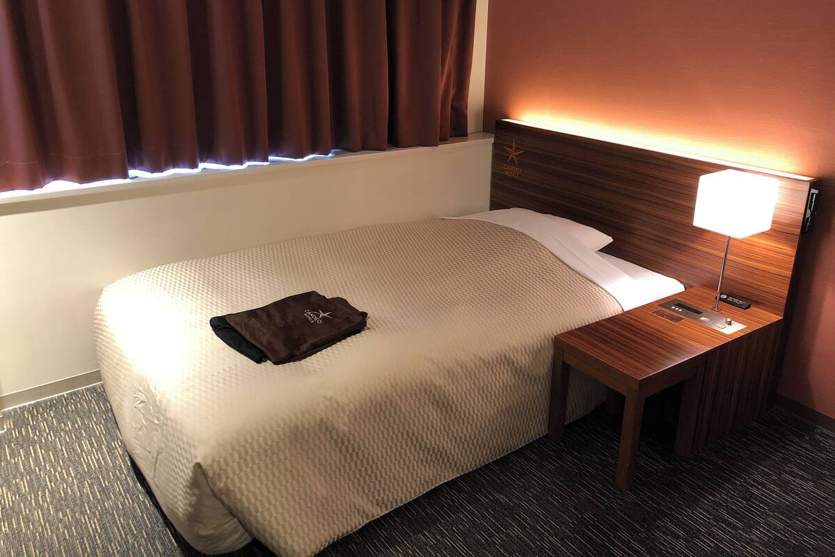 Una habitación de hotel con una cama bien hecha, una lámpara de noche y una ventana con cortinas.