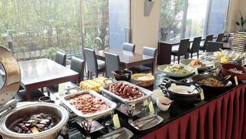 Ein Buffet mit verschiedenen Speisen, darunter Hotdogs, Salate und Desserts, in einem Raum mit großen Fenstern und einem roten Vorhang.