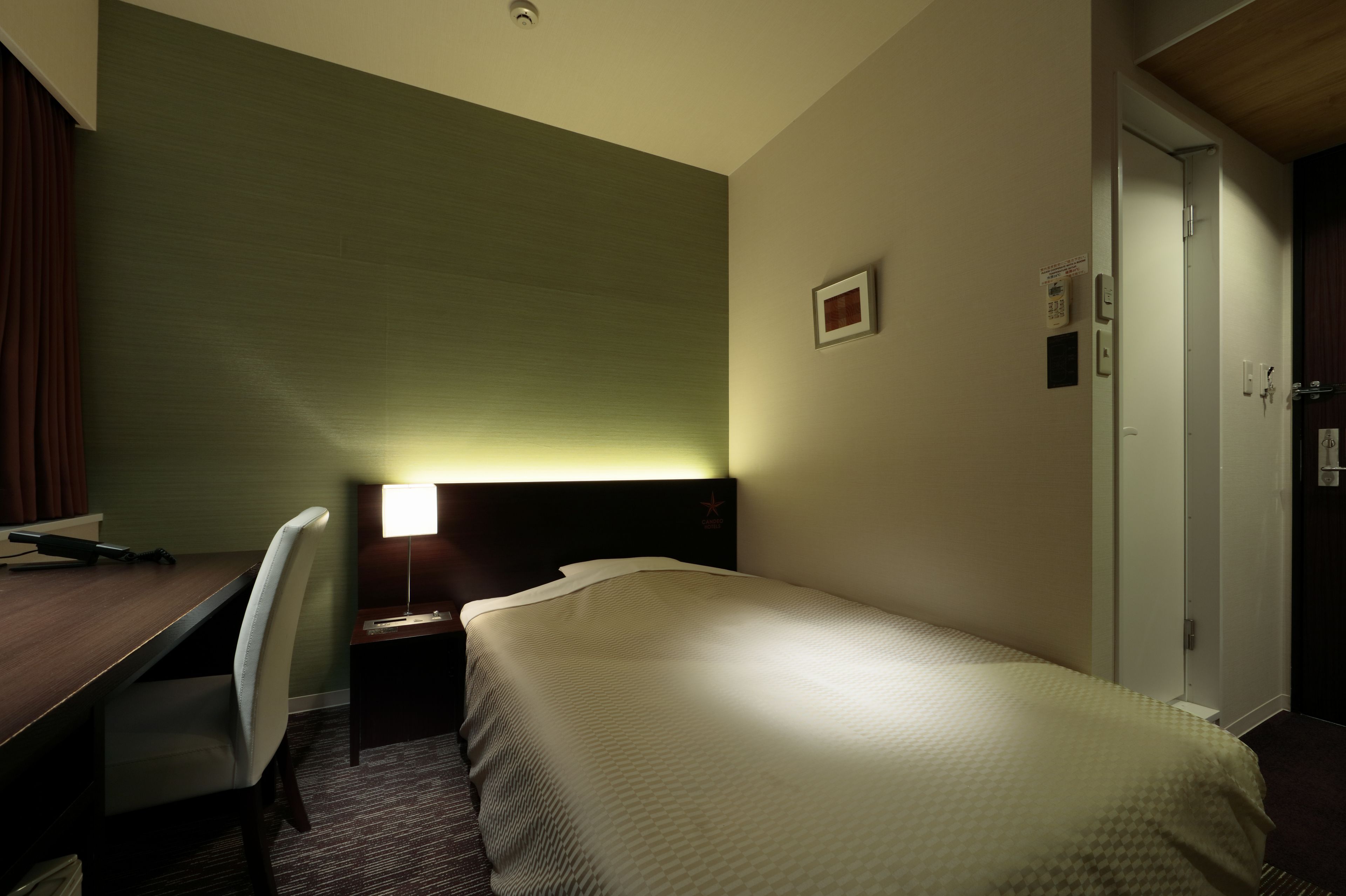 A hotel room with a bed, desk, chair, and a nightstand with a lamp.