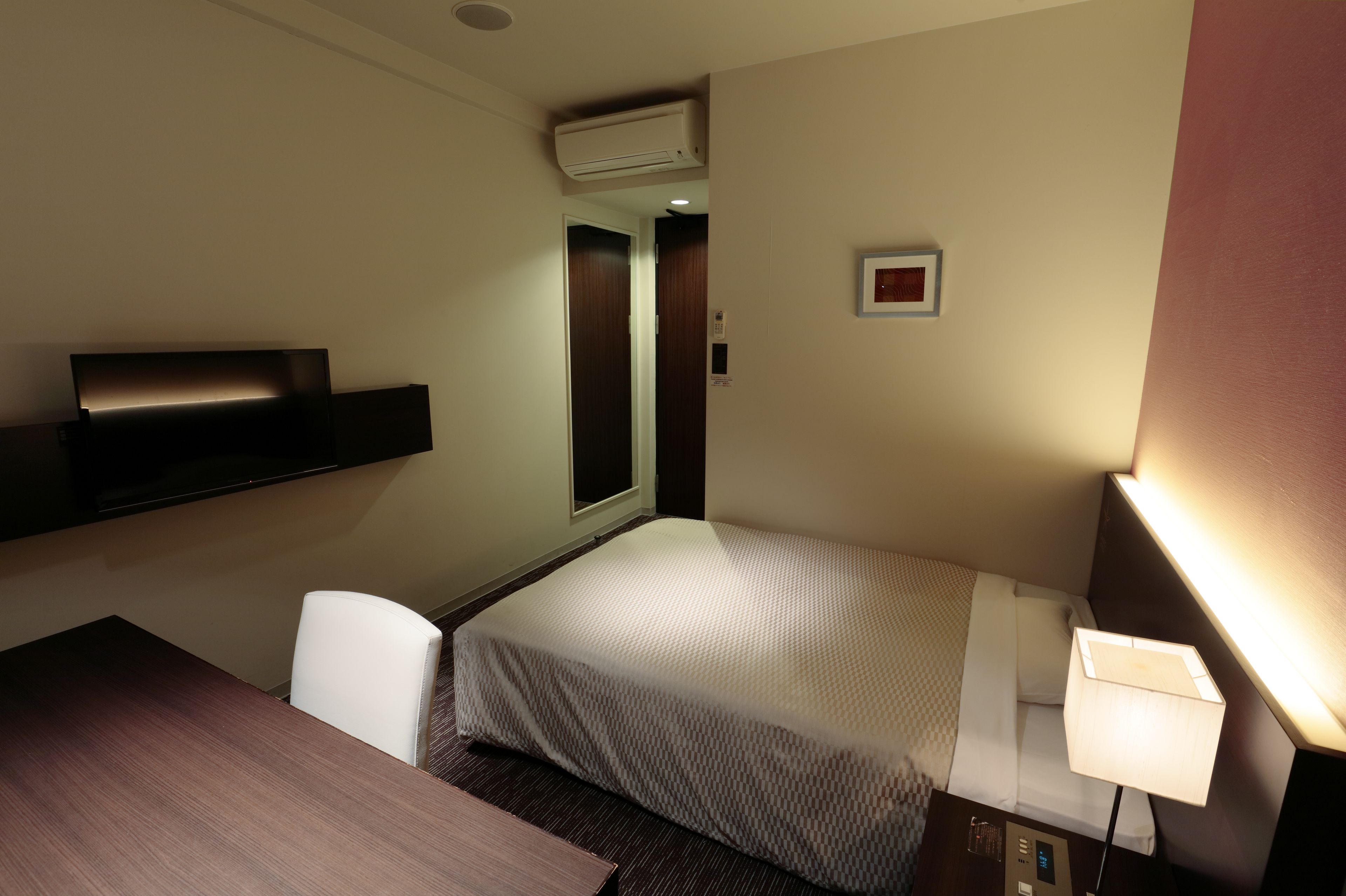 Habitación de hotel pequeña con una cama individual, un escritorio, una silla, una mesita de noche, una lámpara y un equipo de aire acondicionado.