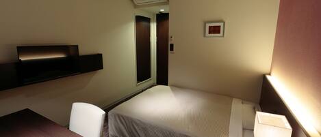 A compact hotel room with a single bed, a desk, a chair, a bedside table, a lamp, and an air conditioning unit.