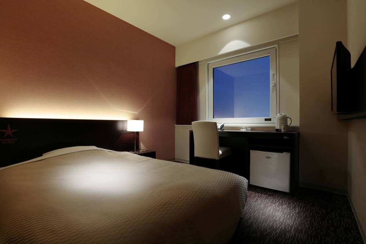 Habitación de hotel con una cama grande, un escritorio con computadora, una silla, un ventanal y un televisor montado en la pared.