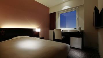 A hotel room with a large bed, a desk with a computer, a chair, a window, and a TV mounted on the wall.
