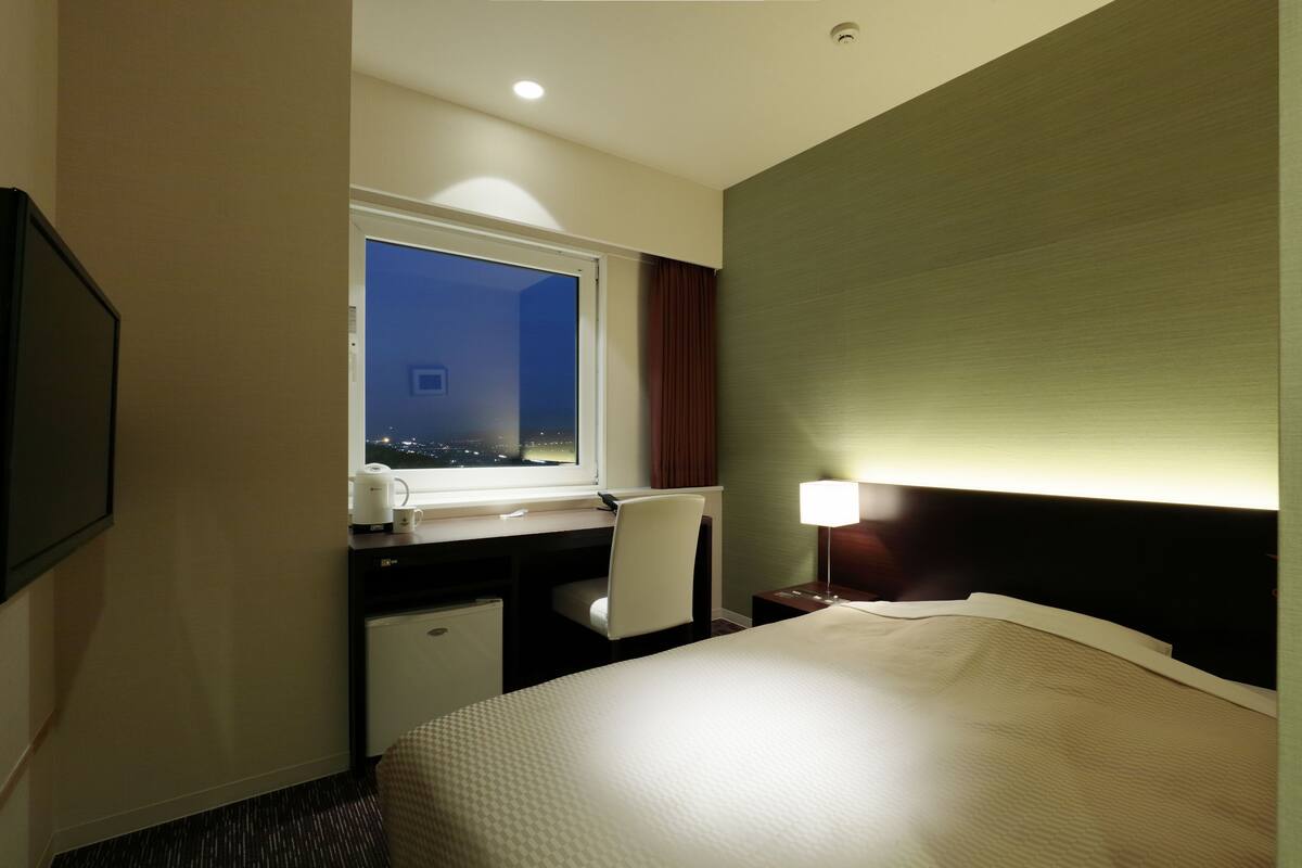 Habitación de hotel con cama, escritorio, silla y ventana con vista a la ciudad de noche.