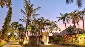 A pathway leading to a building with a thatched roof, surrounded by palm trees and lush greenery. - The Club Villas (Seminyak)