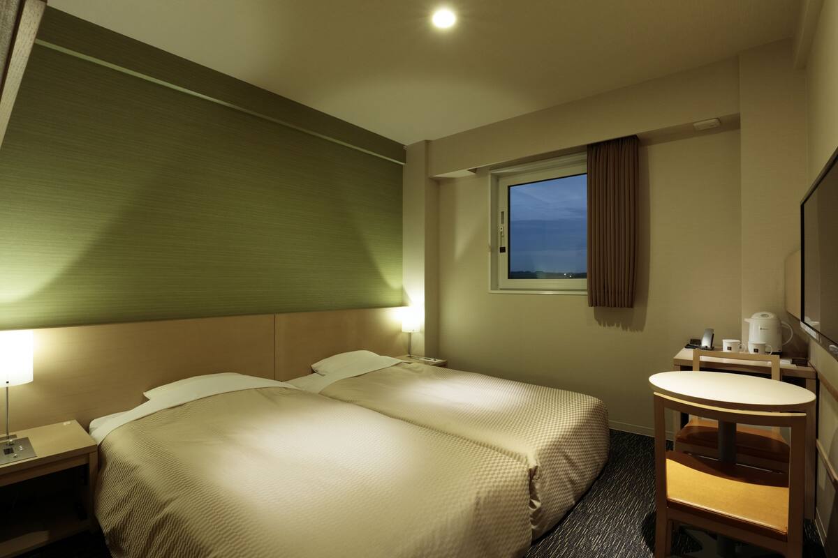 A hotel room with two beds, a small desk with a chair, a window with curtains, and a bathroom with a sink and mirror.