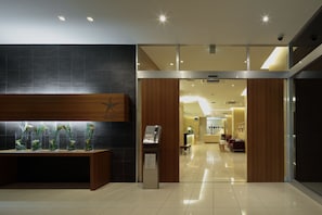 Uma área de recepção de hotel com design moderno, destacando um balcão de atendimento, uma vitrine de flores e vista para o interior do hotel.