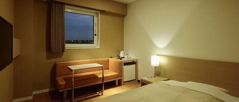 A hotel room with a bed, a desk, a chair, a small table, a nightstand, a lamp, a window with curtains, and a view of the city at dusk.