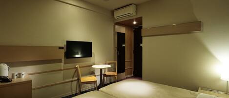 A hotel room with a bed, a desk with a telephone, a small table with chairs, a television mounted on the wall, and an air conditioning unit.