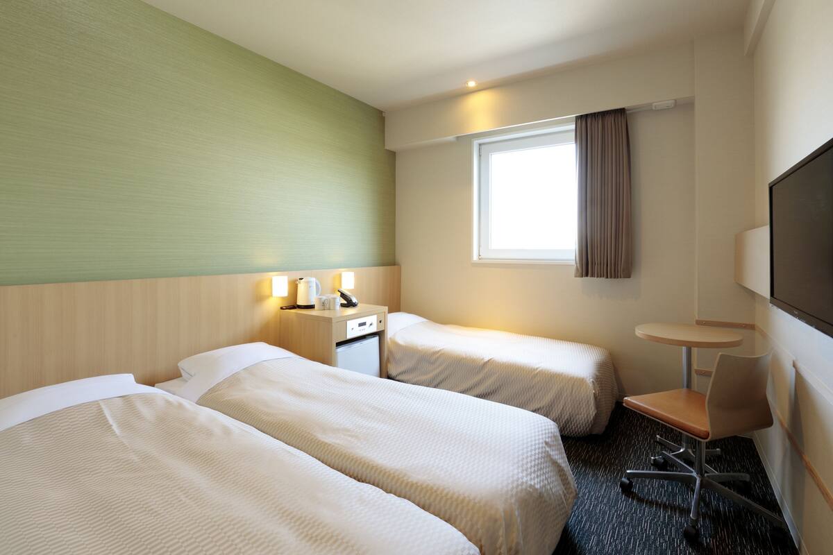 A hotel room with two beds, a small desk, a chair, a TV, and a window with curtains.
