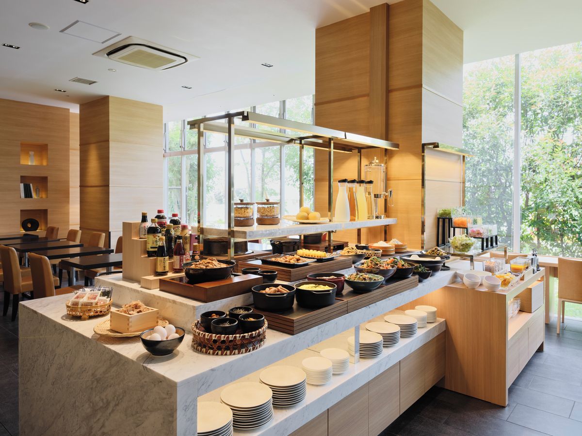 A buffet setup with various food items and condiments, a seating area with tables, and a large window providing natural light.