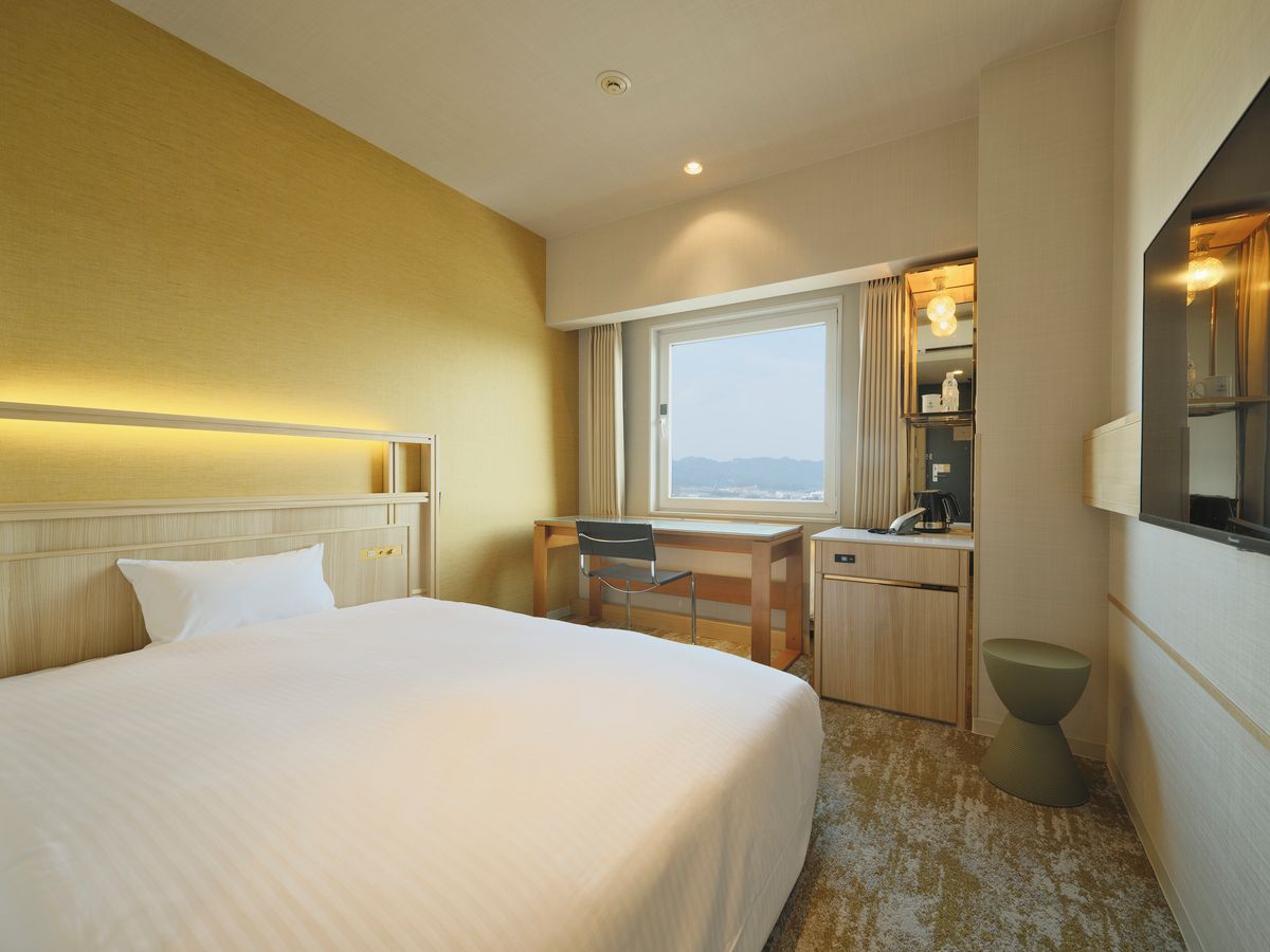 A hotel room with a bed, desk, chair, and a small stool.