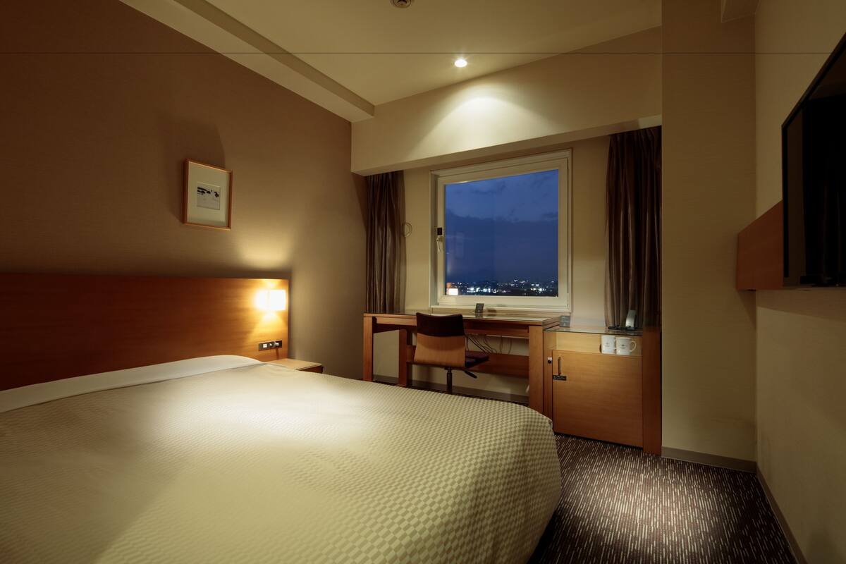 A hotel room with a large bed, a desk with a chair, a window with a view of the city at night, and a framed picture on the wall.