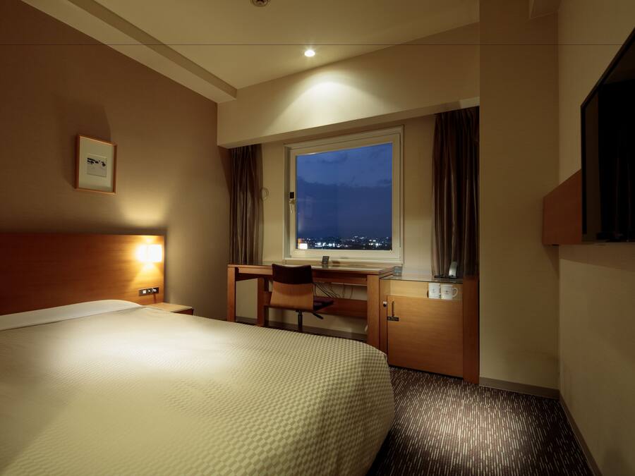 Une chambre d’hôtel avec un grand lit, un bureau avec une chaise, une fenêtre donnant sur la ville la nuit et un tableau encadré au mur.