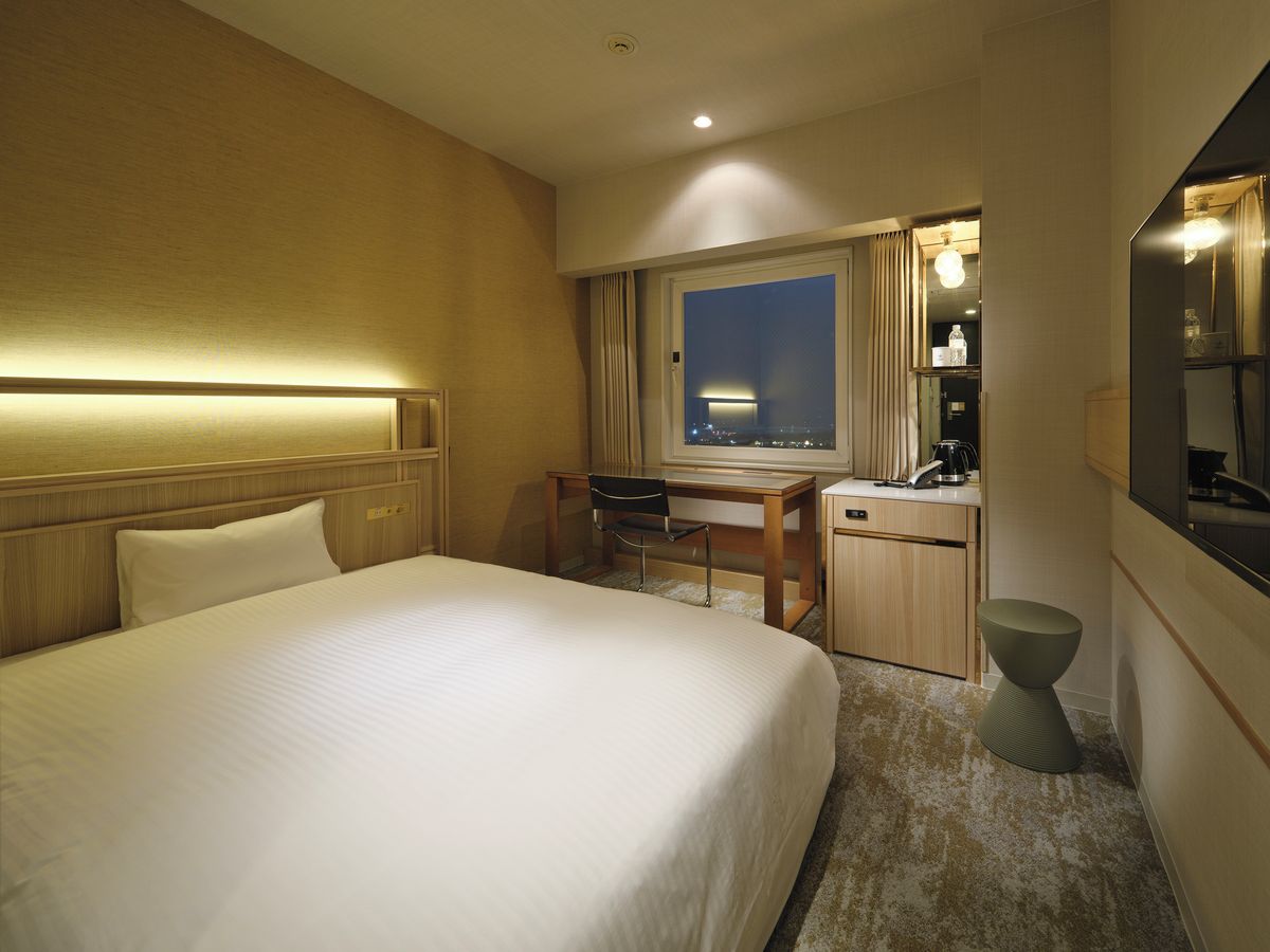 A hotel room with a large bed, a desk with a chair, a small stool, a mirror, and a window with curtains.