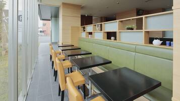 A restaurant with green booths and black tables, a glass wall, and wooden shelves with plants and cups.