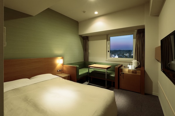 Habitación de hotel con una cama grande, un escritorio con silla, una ventana con cortinas y vistas a la ciudad al anochecer.
