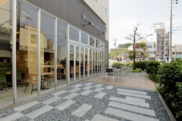 A hotel with a modern glass facade, outdoor seating area, and a paved walkway. - Candeo Hotels Handa (Handa)