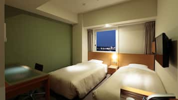 Ein Hotelzimmer mit zwei Betten, einem Schreibtisch, einem Sessel, einem an der Wand befestigten Fernseher und einem Fenster mit Blick auf die beleuchtete Stadt.