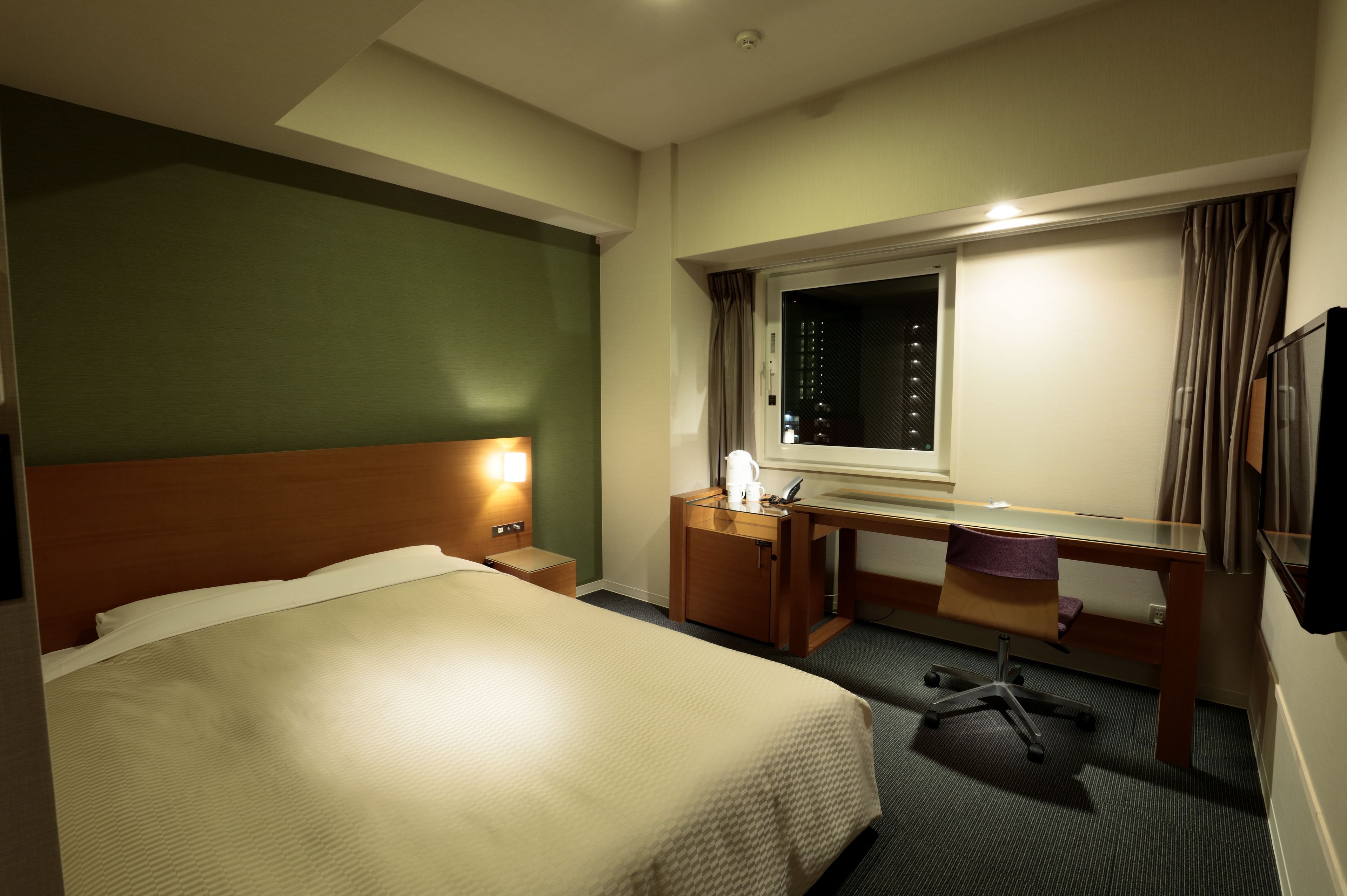 A hotel room with a bed, a desk, a chair, a nightstand, a lamp, and a window with curtains.