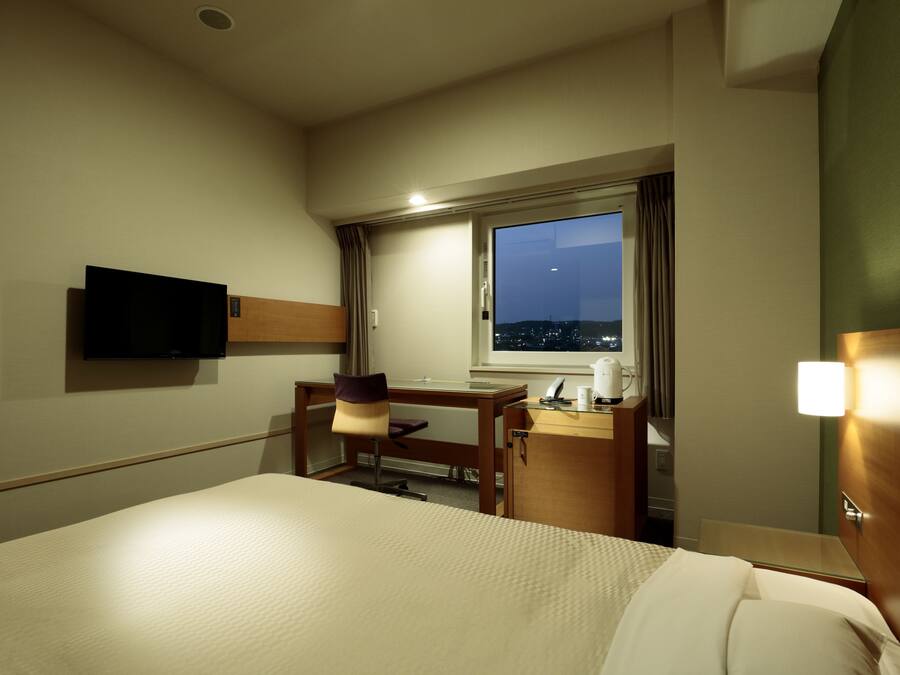 A hotel room with a bed, a desk, a chair, a TV, a window with a night view, and a bedside table.