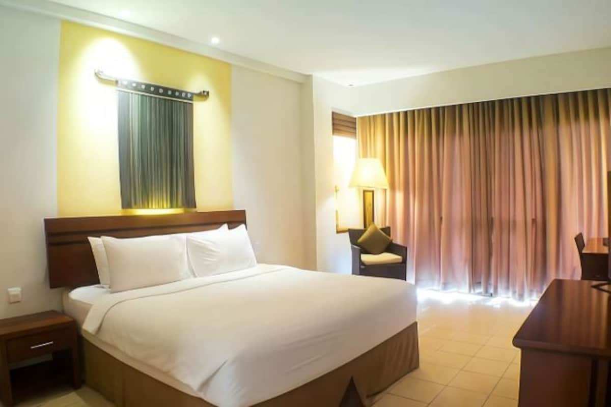 A hotel room with a large bed, a nightstand, a desk, a lamp, and a view of the outside through curtains.