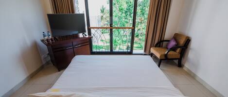 Habitación de hotel con cama, televisor sobre un mueble de madera, una silla con cojín y balcón con vistas a la vegetación.