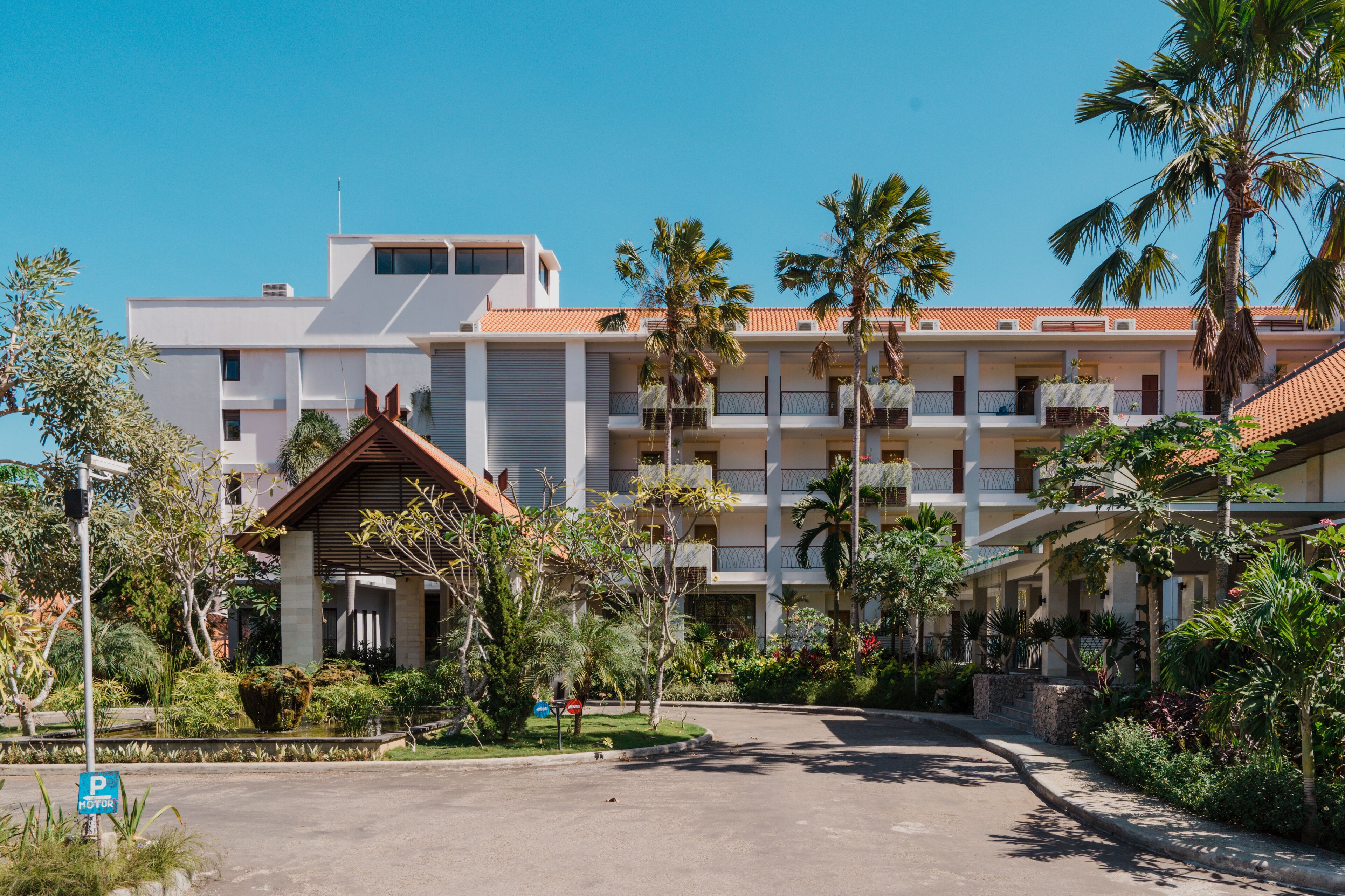 Un hotel de varios pisos con un patio central, palmeras y un estacionamiento.