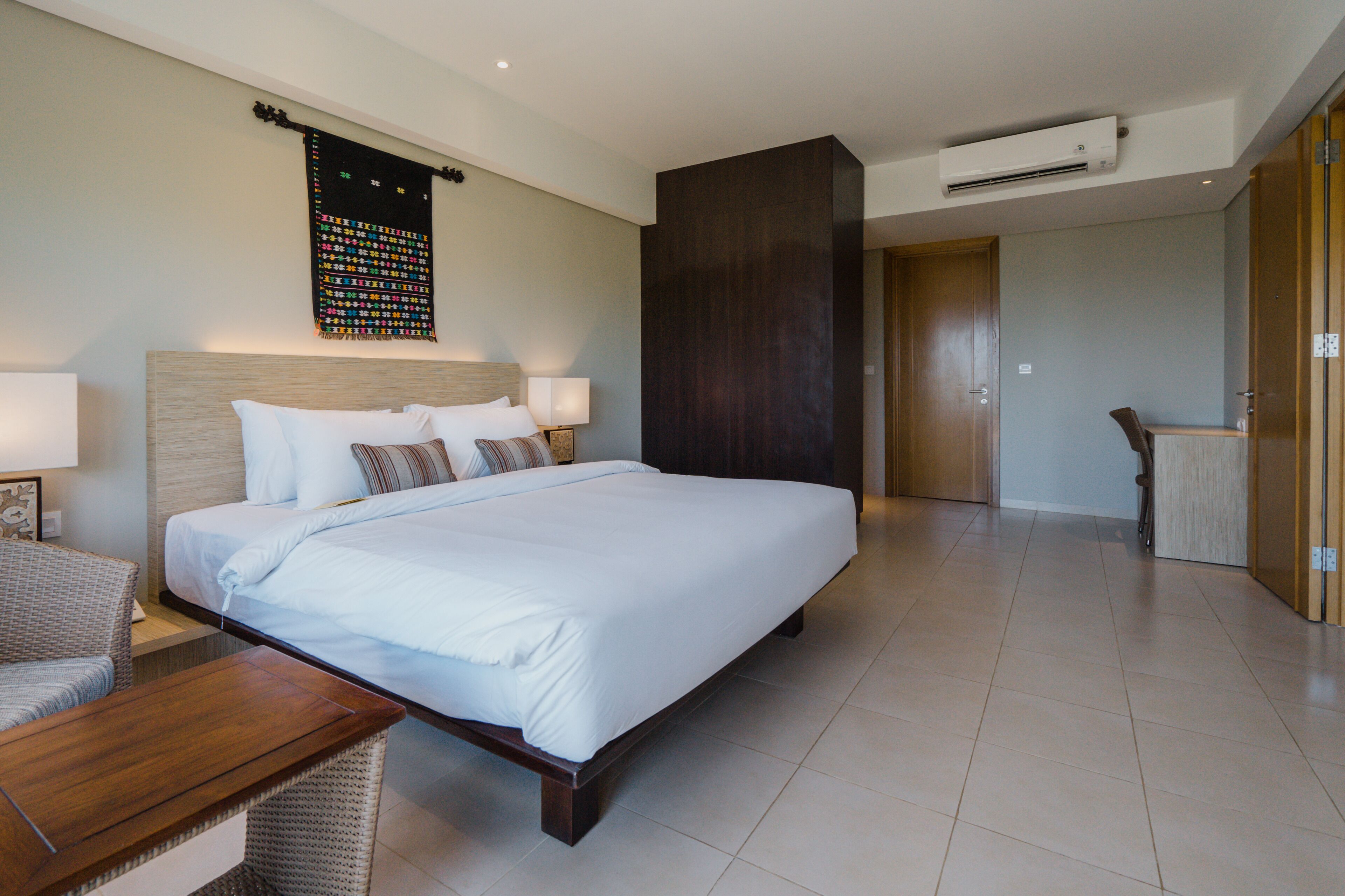 Ruime hotelkamer met een groot bed, een houten nachtkastje, een stoel en een wandairconditioner.