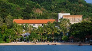 Ett hotell vid stranden med en byggnad som har rött tak, palmer och utsikt över havet.