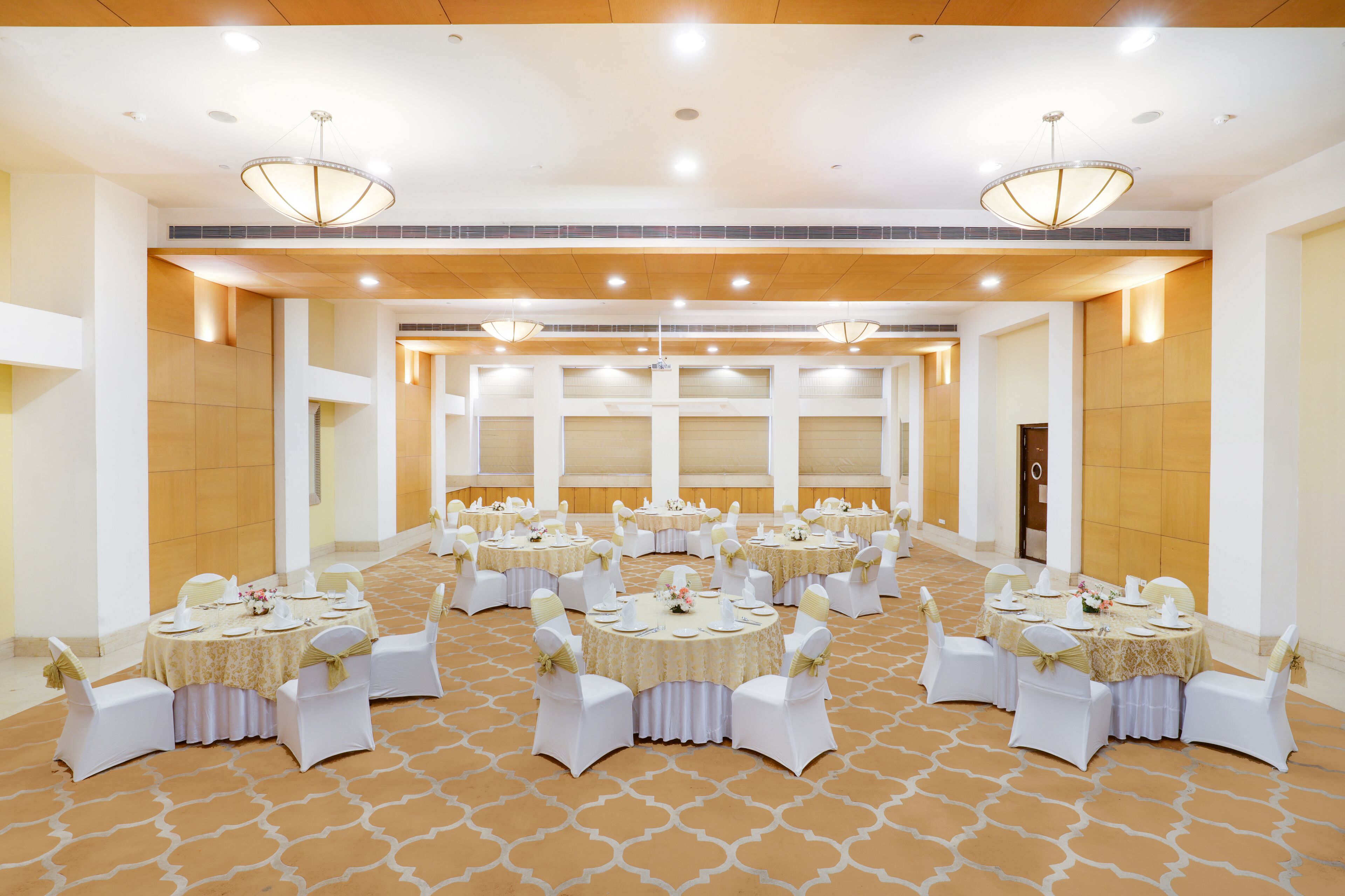 Un salón de banquetes con varias mesas redondas, cada una con sillas blancas y manteles, y un piso con diseño.