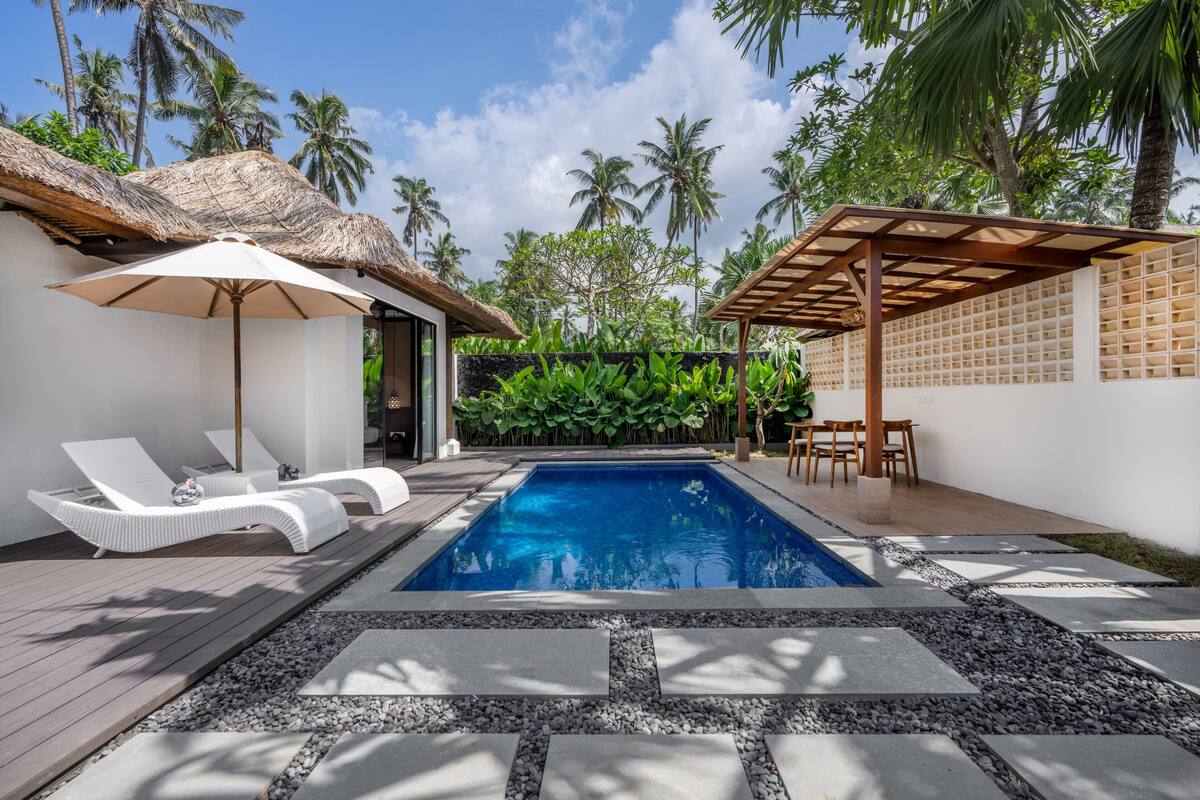 A swimming pool with lounge chairs and umbrellas, surrounded by palm trees and a thatched roof structure.