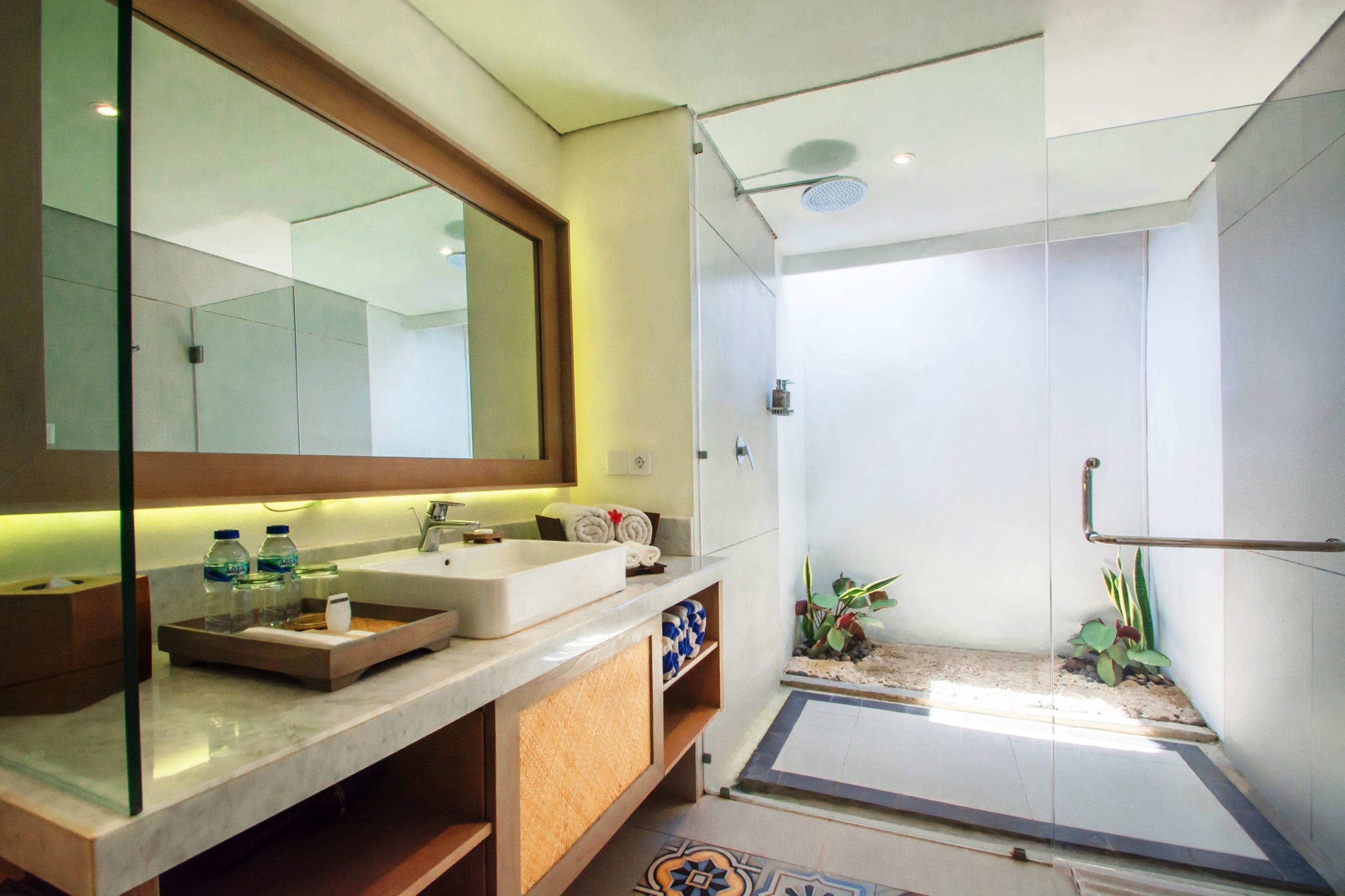 A modern bathroom with a large mirror, two sinks, a shower area, and a small plant.
