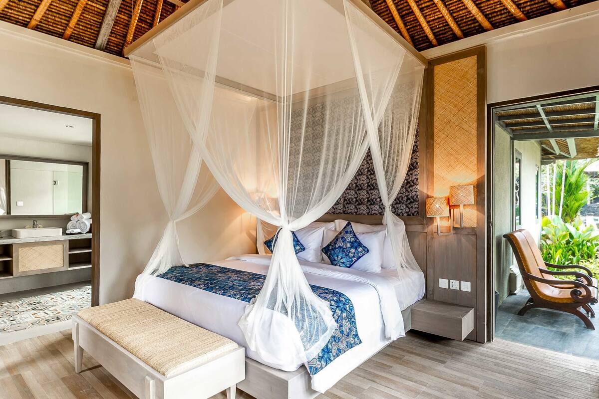 A bedroom with a canopy bed, bedside table, bench, and a view of the outdoors.