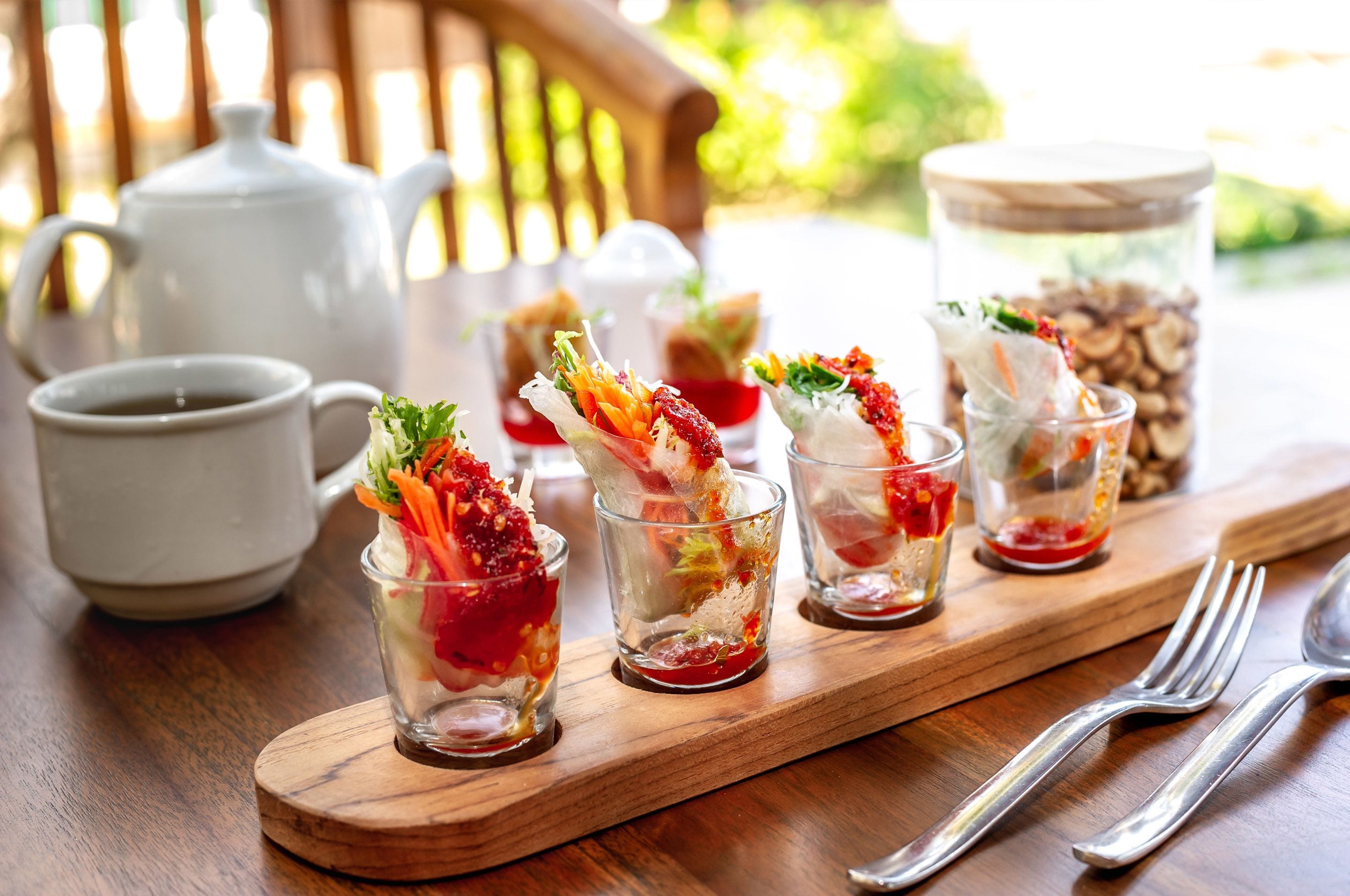 A wooden table with a tea set, including a teapot and cups, and a glass tray holding four small glasses with colorful food garnishes.