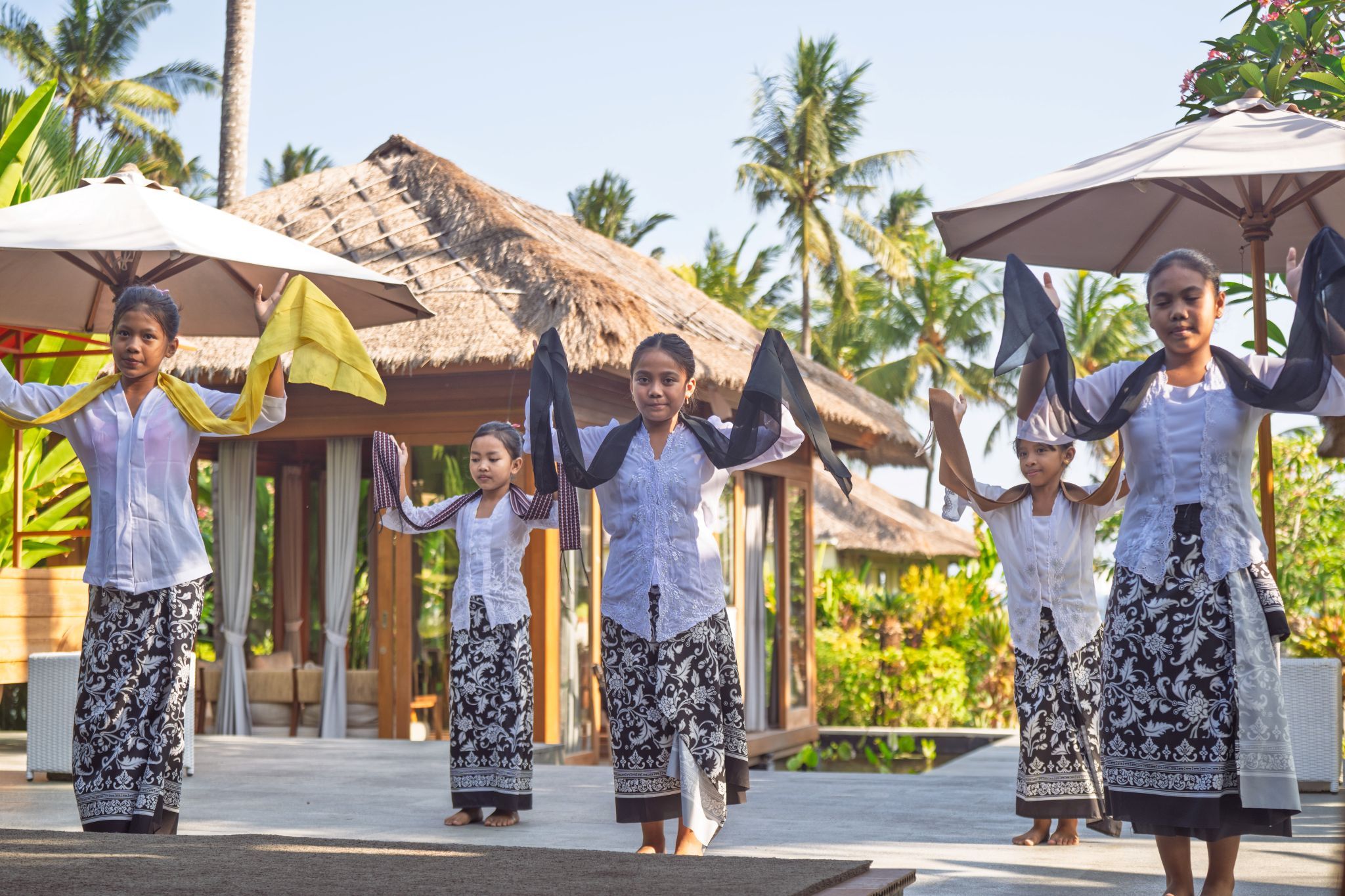 A group of people in traditional attire performing a dance in an outdoor setting with thatched-roof structures and palm trees in the background.