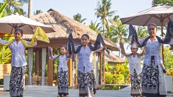 A group of people in traditional attire performing a dance in an outdoor setting with thatched-roof structures and palm trees in the background.