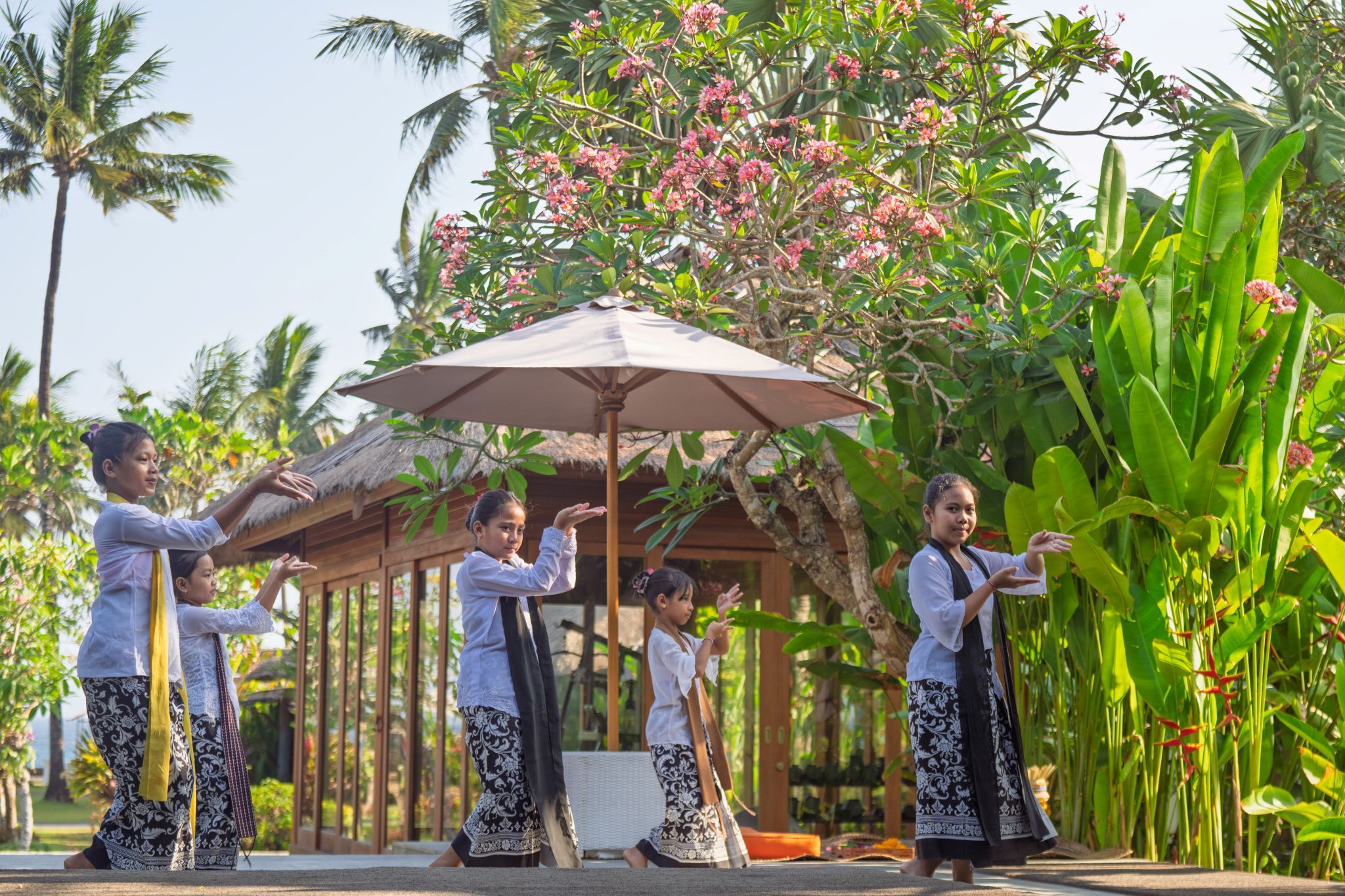 A group of people performing a traditional dance in an outdoor setting with a thatched-roof pavilion and lush tropical vegetation.