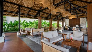 A spacious indoor area with wooden furniture, a thatched roof, and a view of lush greenery outside. - Nirwana Beach & Resort (Manggis)
