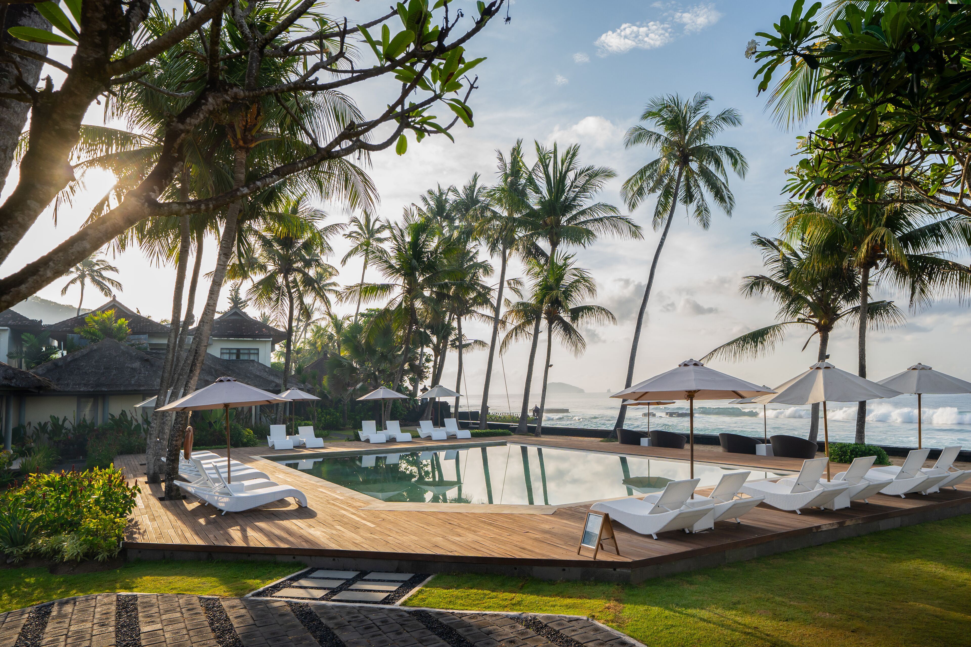 A poolside area with lounge chairs and umbrellas, surrounded by palm trees and overlooking the ocean.