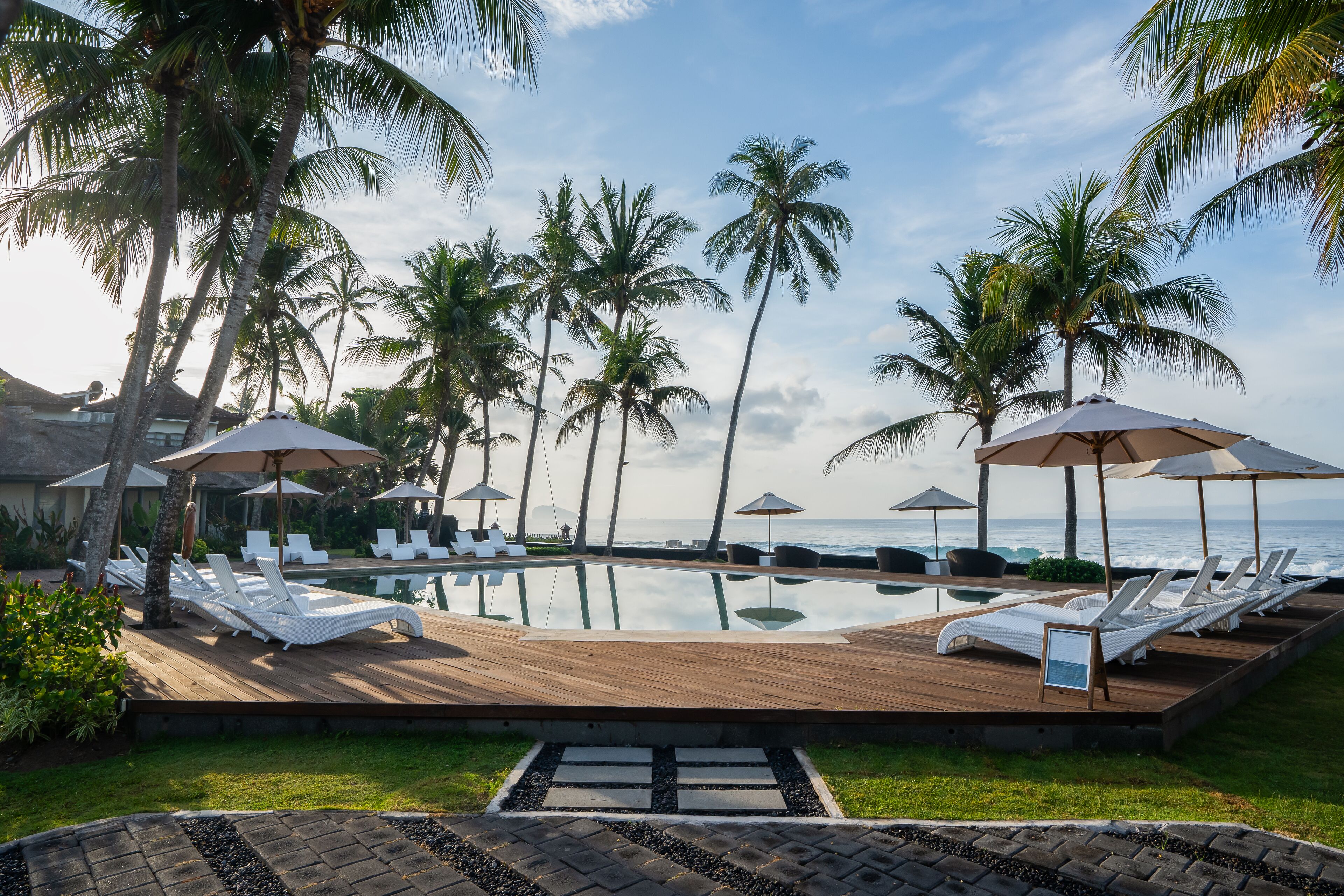 A poolside area with lounge chairs and umbrellas, surrounded by palm trees and overlooking the ocean.