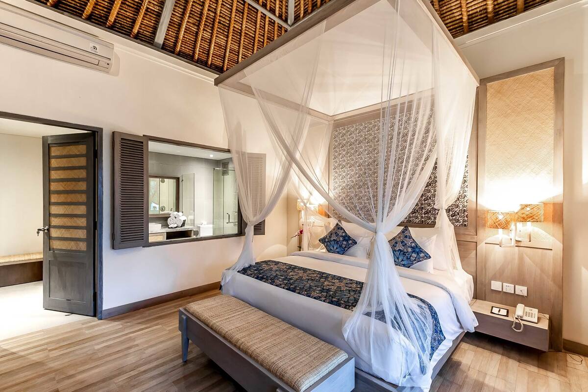 A bedroom with a four-poster bed, a bedside table with a lamp, a bench, and a large mirror.