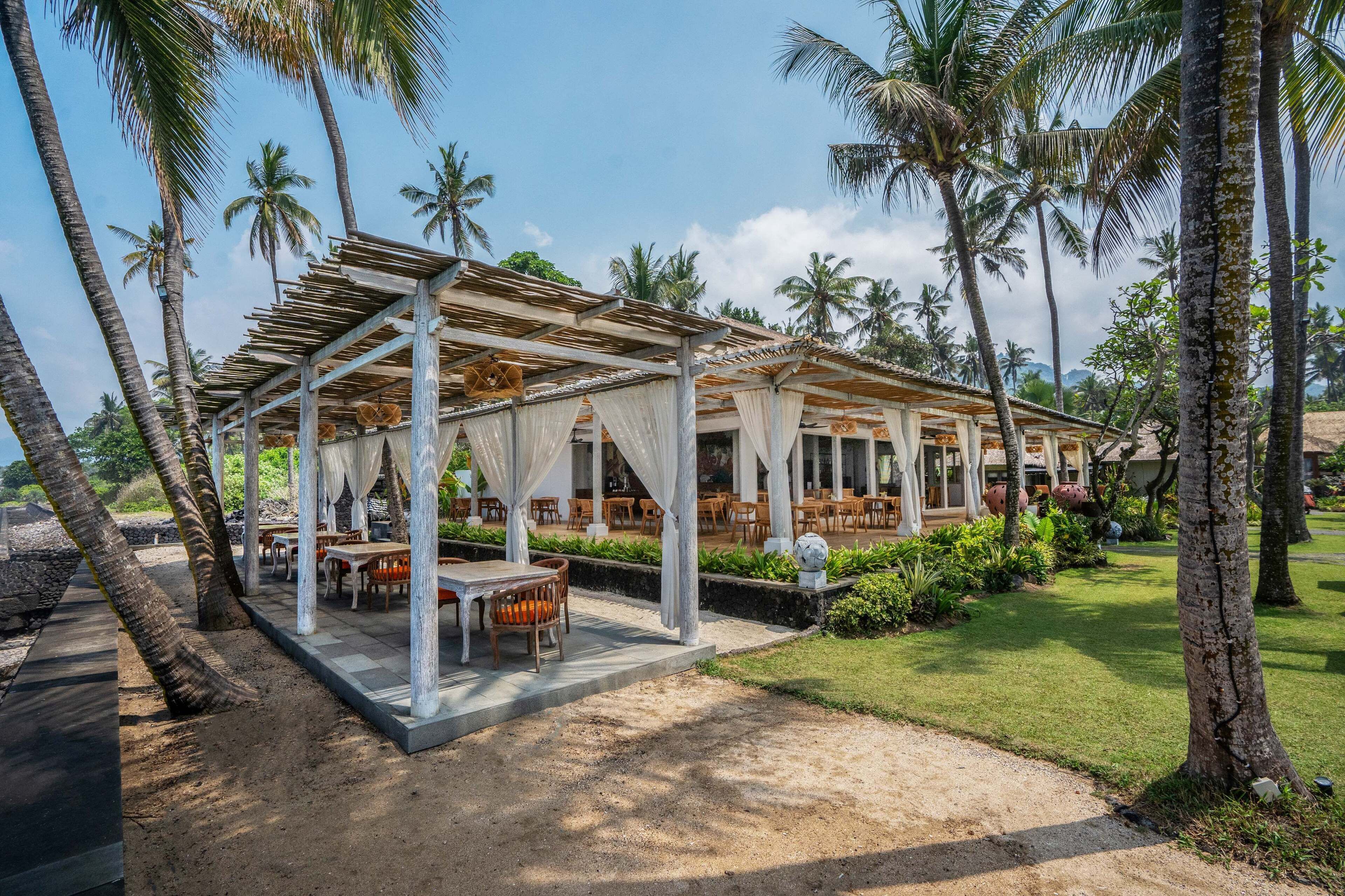 A covered outdoor dining area with tables and chairs, surrounded by palm trees and greenery.