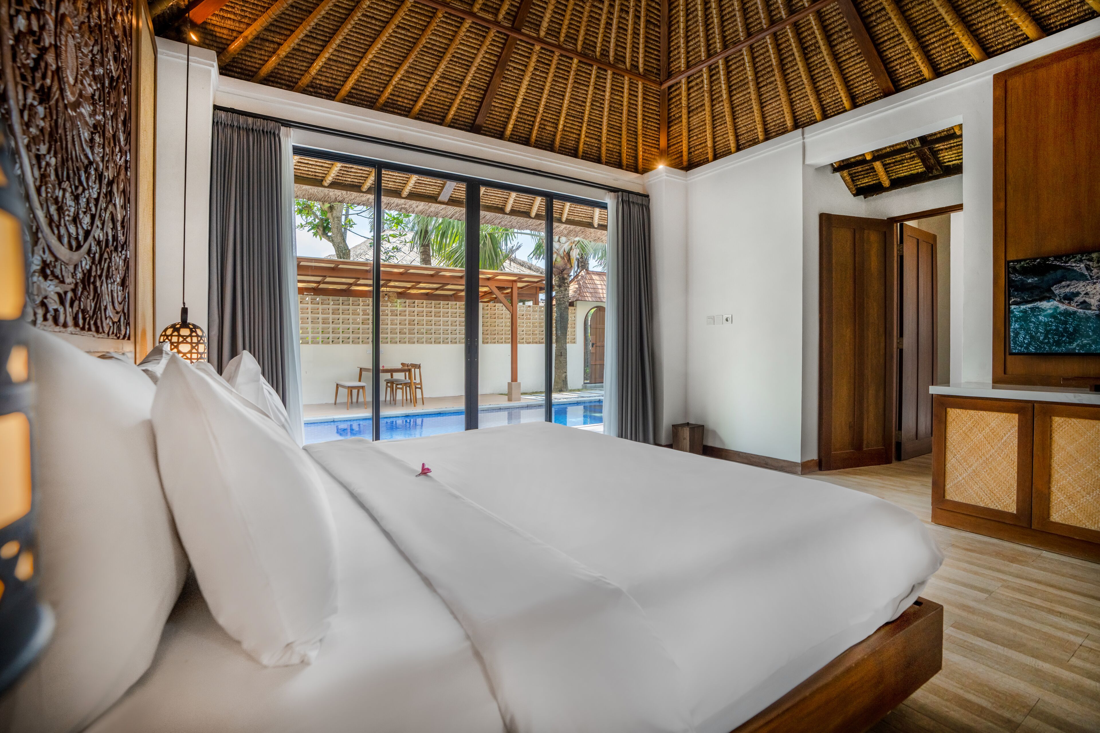 A bedroom with a large bed, a TV, a wooden desk, and a view of a pool.