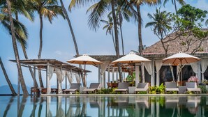 A poolside area with lounge chairs and umbrellas, surrounded by palm trees and a clear view of the ocean. - Nirwana Beach & Resort (Manggis)
