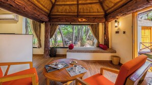 Una habitación acogedora con techo de madera, una ventana con vista a árboles, una mesita con una taza y una revista, y sillones color naranja.