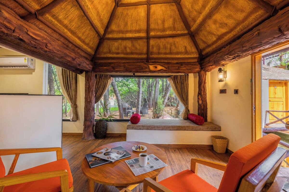 A cozy room with a wooden ceiling, a window with a view of trees, a small table with a cup and a magazine, and orange chairs.