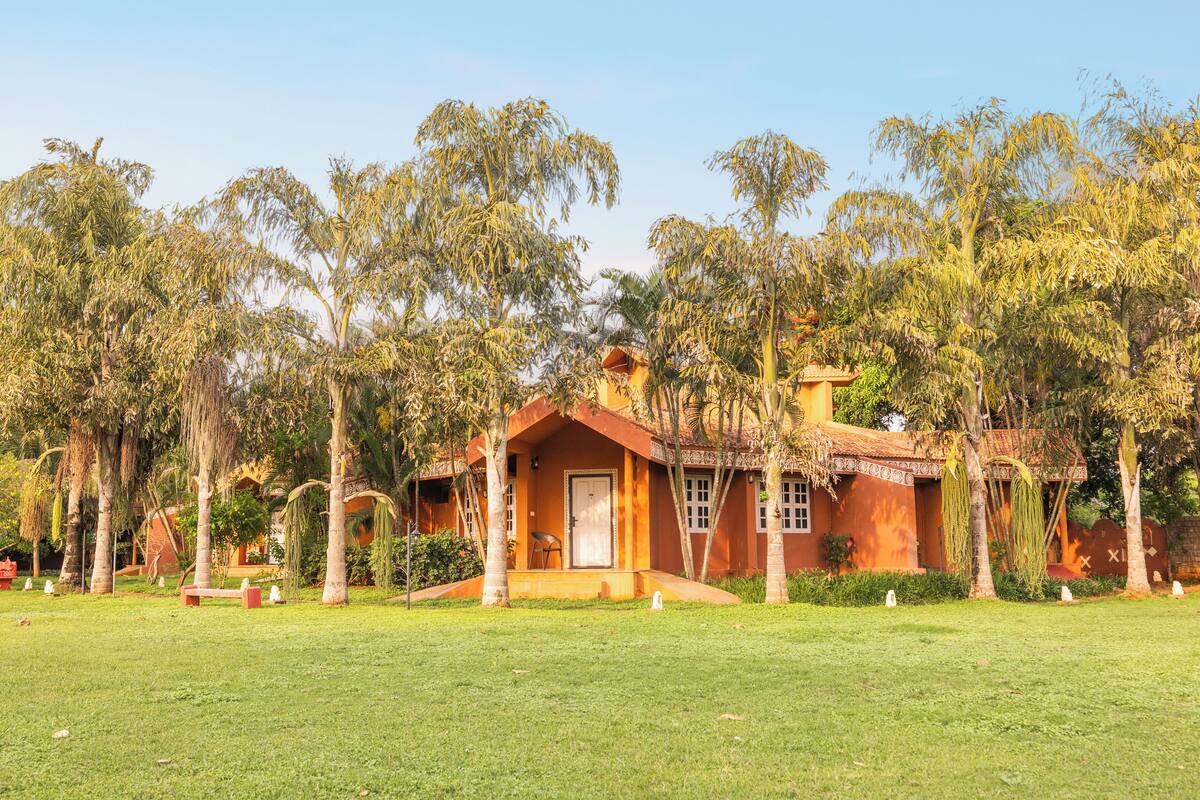A small, single-story house with a red roof and beige walls, surrounded by a well-maintained lawn and tall trees.